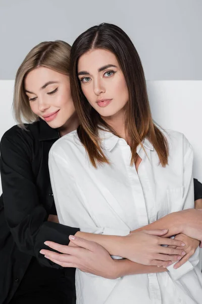 Smiling blonde woman embracing brunette friend looking at camera on white and grey background — Stock Photo