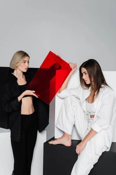 Blonde woman holding red glass near barefoot friend and black and white cubes isolated on grey — Stock Photo