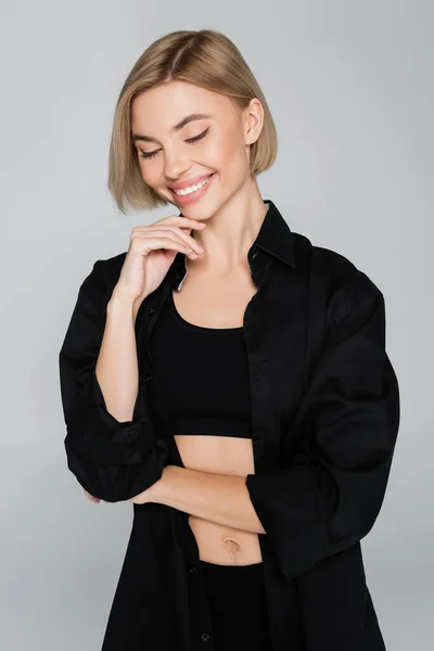 Bonita mujer en sujetador negro y camisa tocando la barbilla y sonriendo aislado en gris - foto de stock