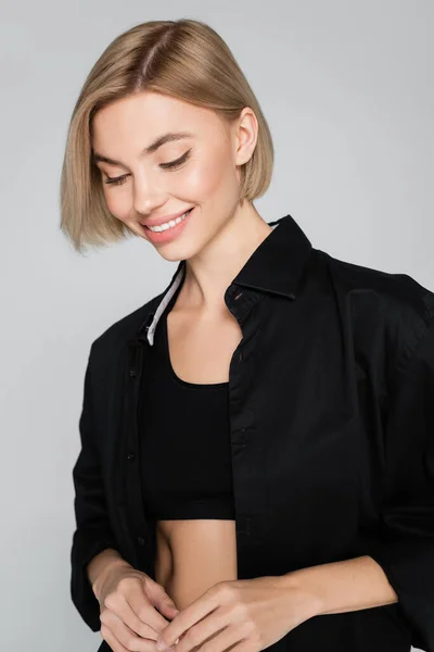 Young blonde woman in black bra and shirt smiling isolated on grey — Stock Photo