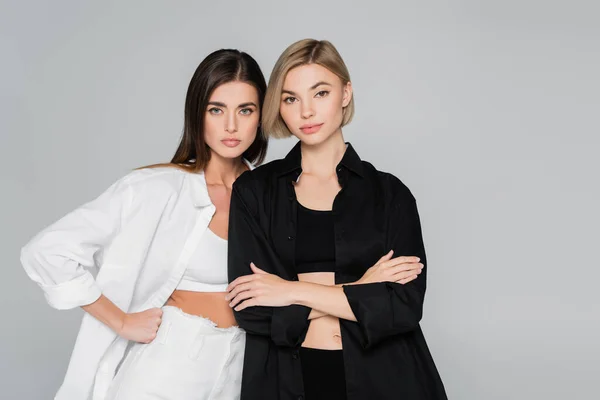 Femmes en chemises noires et blanches debout avec la main sur la hanche et les bras croisés isolés sur le gris — Photo de stock