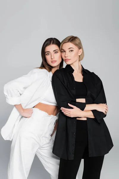 Mujeres en ropa blanca y negra posando con los brazos cruzados y la mano en la cadera aislada en gris - foto de stock