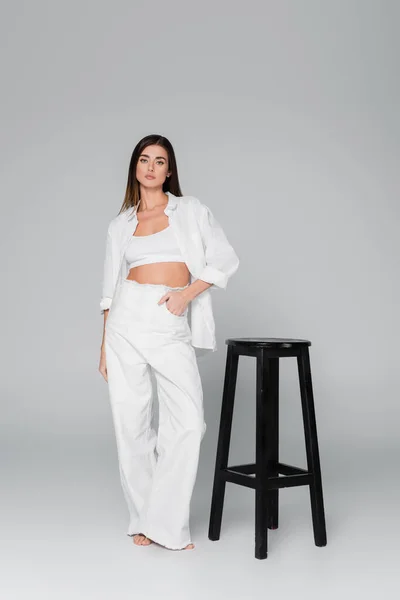 Full length of woman standing with hand in pocket of white trousers near black stool on grey background — Stock Photo