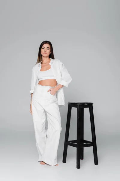 Toute la longueur de la femme debout avec la main dans la poche de pantalon blanc près de tabouret noir sur fond gris — Photo de stock