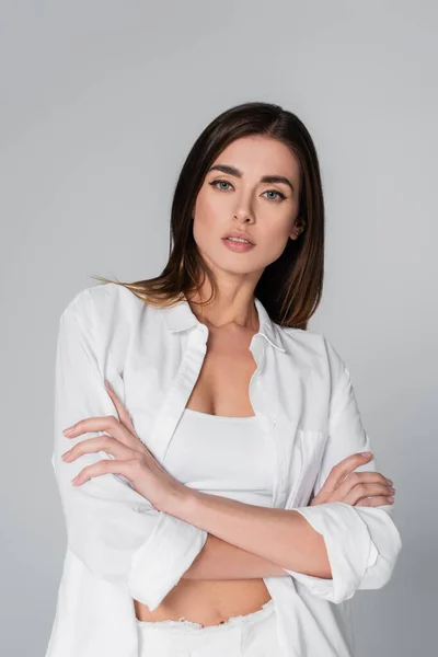 Brunette woman in white shirt crossing arms and looking at camera isolated on grey — Stock Photo