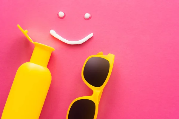 Вид сверху солнцезащитных очков рядом со знаком улыбки и солнцезащитным кремом на розовом фоне — стоковое фото