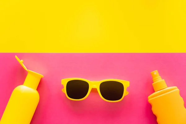 Вид сверху солнцезащитных очков рядом с бутылками солнцезащитных кремов на желтом и розовом фоне — стоковое фото