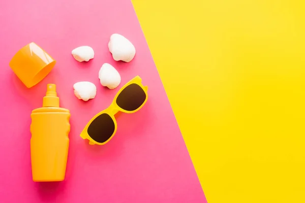Vista superior de protetor solar e óculos de sol perto de conchas no fundo rosa e amarelo — Fotografia de Stock