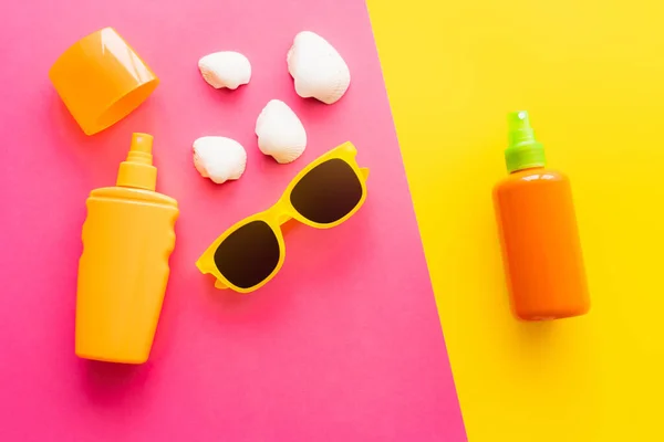 Vista superior do protetor solar em garrafas perto de óculos de sol e conchas no fundo rosa e amarelo — Fotografia de Stock