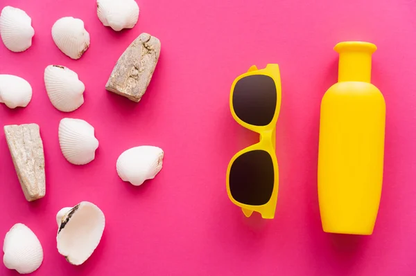 Vista superior de conchas marinas cerca de gafas de sol y botella de protector solar sobre fondo rosa - foto de stock