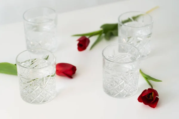 Facettierte Gläser mit Wasser in der Nähe roter Tulpen auf weißem, verschwommenem Hintergrund — Stockfoto