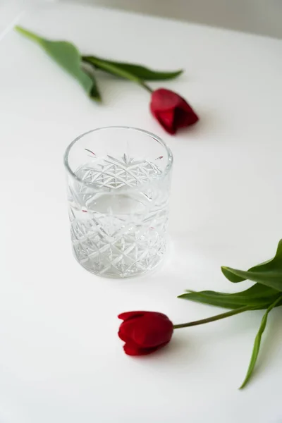 Vidrio con agua pura cerca de tulipanes rojos frescos en la superficie blanca y fondo borroso - foto de stock