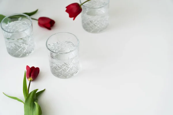 Óculos com padrão facetado e água limpa perto de tulipas vermelhas na superfície branca — Fotografia de Stock