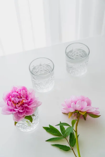 Vue grand angle des verres à facettes avec de l'eau et des pivoines roses sur la surface blanche — Photo de stock