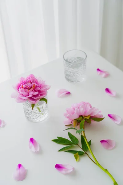 Visão de alto ângulo de copos com água e peônias rosa na mesa branca — Fotografia de Stock