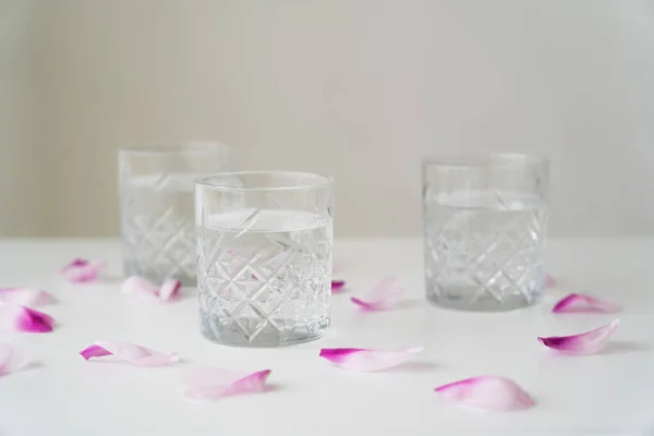 Blütenblätter und facettierte Gläser mit reinem Wasser auf weißer Tischplatte isoliert auf grau — Stockfoto