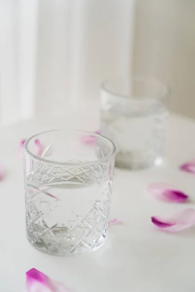 Vista da vicino di vetro sfaccettato con acqua pulita vicino petali floreali su sfondo grigio sfocato — Foto stock