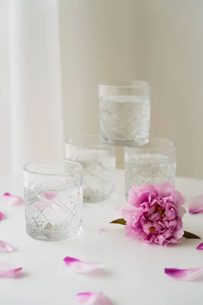 Vidrios facetados apilados cerca de peonía rosa y pétalos sobre mesa blanca y fondo gris - foto de stock