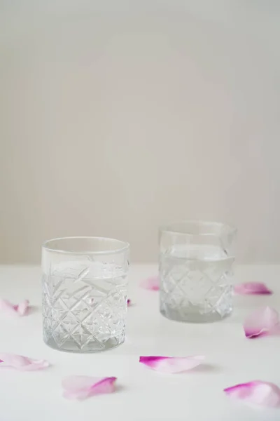 Verres transparents à l'eau douce près des pétales floraux sur plateau blanc isolé sur gris — Photo de stock