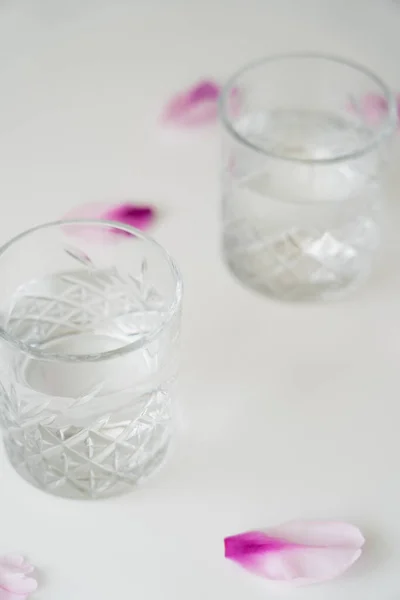 Vue rapprochée des verres avec de l'eau douce près des pétales floraux sur fond blanc et flou — Photo de stock