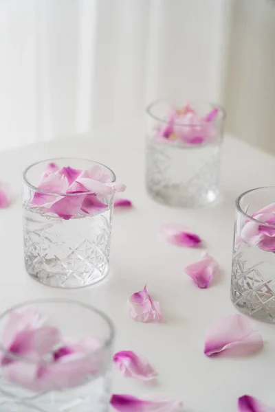 Verres en cristal avec des pétales floraux toniques et naturels sur plateau blanc et fond flou — Photo de stock