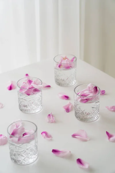 Faceted glasses with gin tonic near floral petals scattered on white tabletop on grey background — Stock Photo