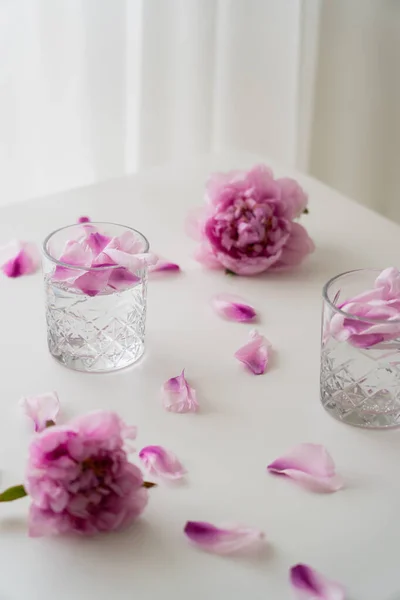 Peonie fresche e petali vicino a bicchieri con gin tonic su tavolo bianco e sfondo grigio — Foto stock