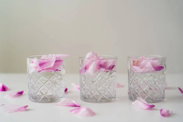 Crystal glasses with floral petals and tonic on white tabletop isolated on grey — Stock Photo