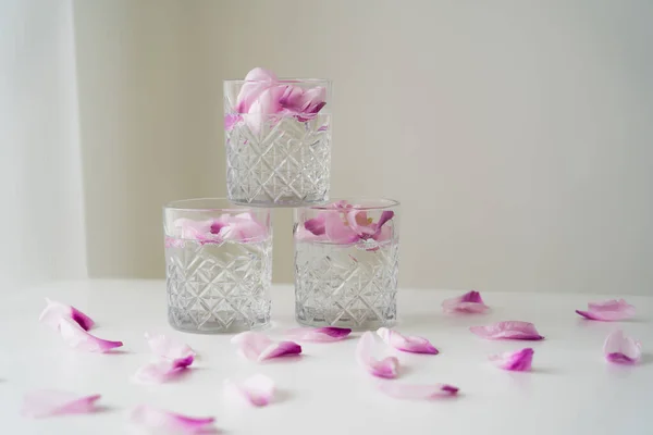 Copos de cristal com pétalas tônicas e florais na superfície branca e fundo cinza — Fotografia de Stock