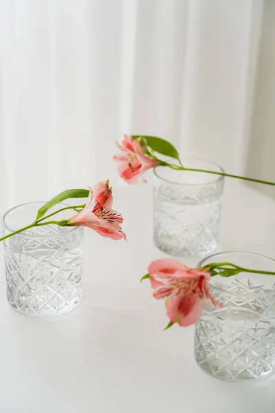 Crystal glasses with water near alstroemeria flowers on white tabletop and grey background — Stock Photo