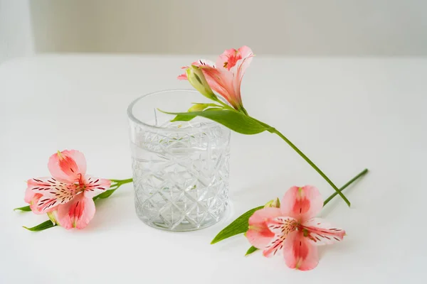 Fiori alstroemeria vicino vetro sfaccettato con acqua pulita su superficie bianca e sfondo grigio — Foto stock