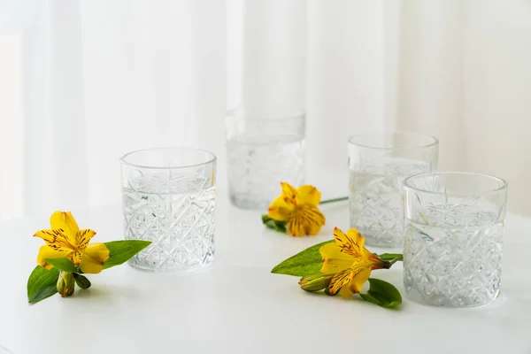 Gelbe Alstroemeria-Blüten in der Nähe facettierter Gläser mit klarem Wasser auf weißem Hintergrund — Stockfoto