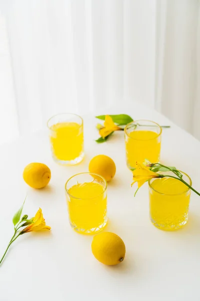 Glasses with lemonade near fresh lemons and yellow alstroemeria flowers on white tabletop — Stock Photo