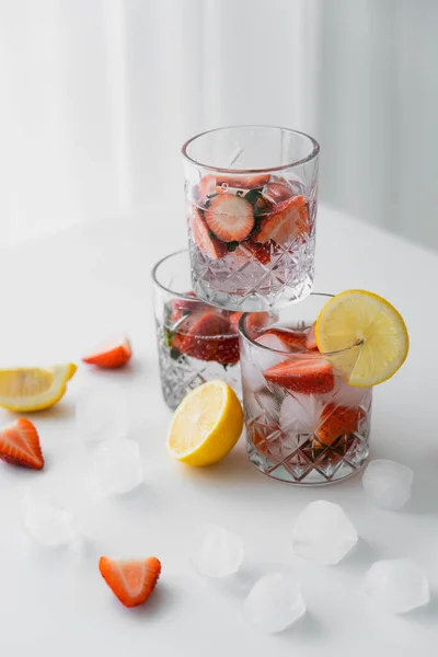 Vasos con tónico de fresa fría cerca de limones cortados y cubitos de hielo en la mesa blanca - foto de stock