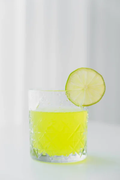 Faceted glass with natural lemonade and slice of lime on grey background — Stock Photo