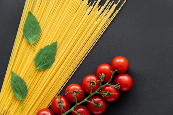 Vue du dessus des tomates cerises près des spaghettis crus et du basilic sur fond noir — Photo de stock