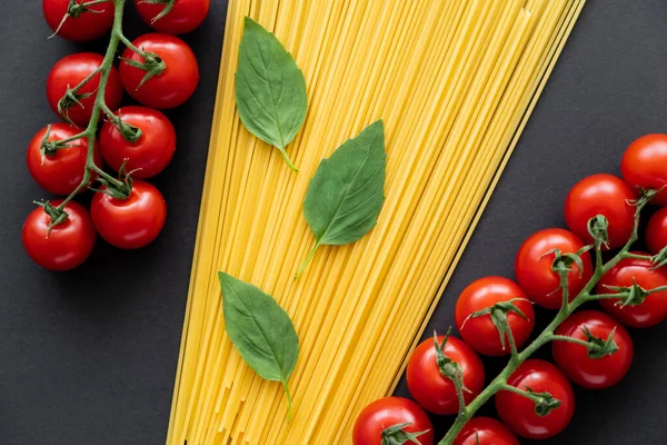 Top view of fresh basil on raw pasta near cherry tomatoes on black background — Stock Photo