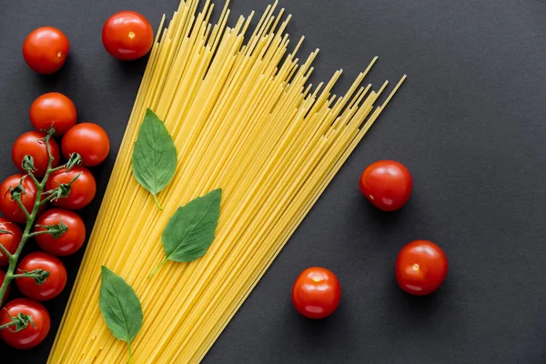 Top view of whole cherry tomatoes near fresh basil and raw spaghetti on black background — Stock Photo