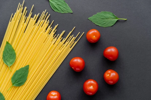 Fresh basil laves near raw pasta and cherry tomatoes on black background — Stock Photo