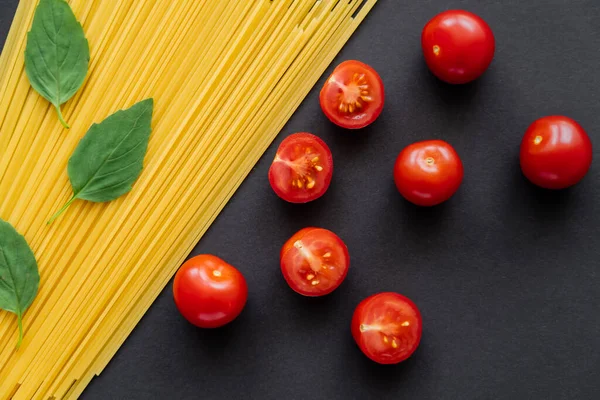 Vue du dessus des tomates cerises coupées et du basilic frais sur des spaghettis crus sur fond noir — Photo de stock