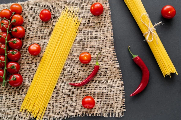 Vista dall'alto di spaghetti crudi vicino a pomodorini e peperoncini su sacco su sfondo nero — Foto stock