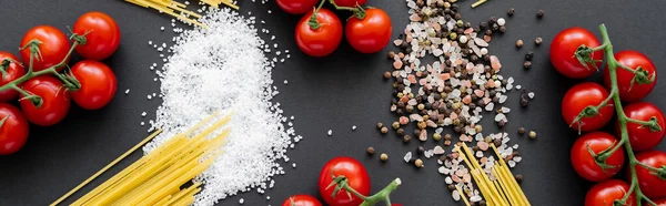 Flat lay with uncooked pasta and cherry tomatoes near spices on black background, banner — Stock Photo