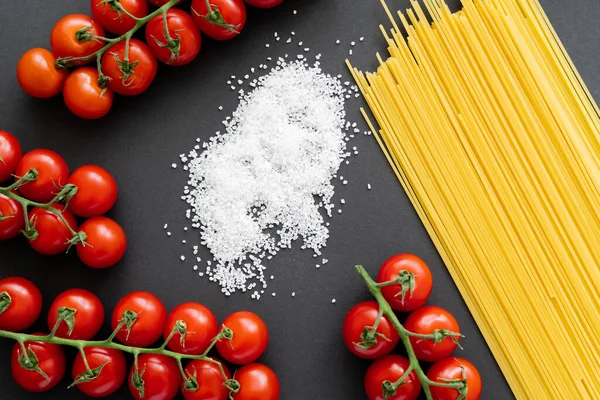 Вид сверху на соль рядом с сырыми спагетти и вишневыми помидорами на черном фоне — стоковое фото