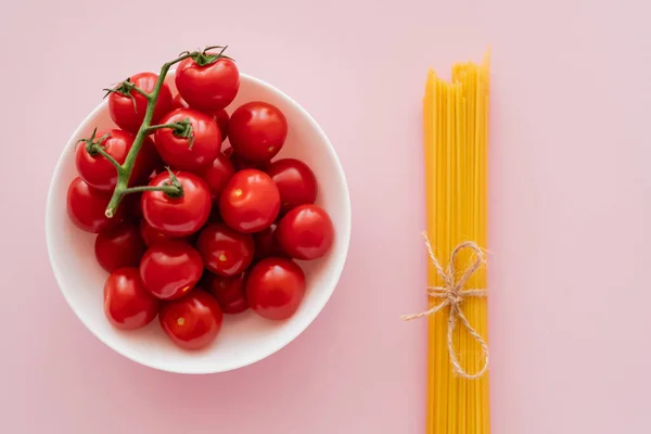 Вид зверху на вишневі помідори в мисці і неварені спагетті на рожевій поверхні — стокове фото