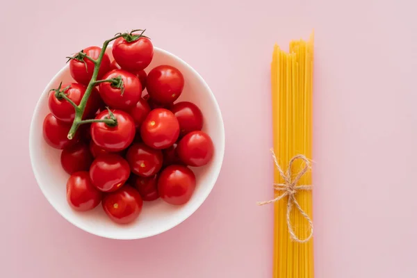 Vista dall'alto di pomodorini ciliegini in ciotola e spaghetti crudi sulla superficie rosa — Foto stock