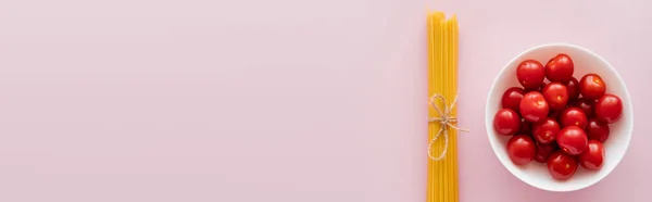 Top view of raw spaghetti and cherry tomatoes in bowl on pink surface, banner — Stock Photo