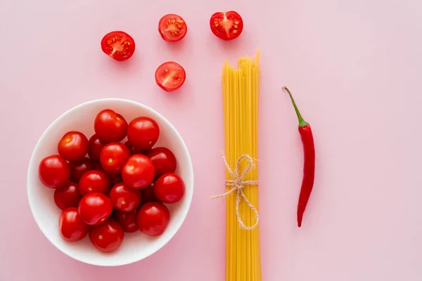 Top view of cherry tomatoes in bowl near raw pasta and chili pepper on pink surface — Stock Photo