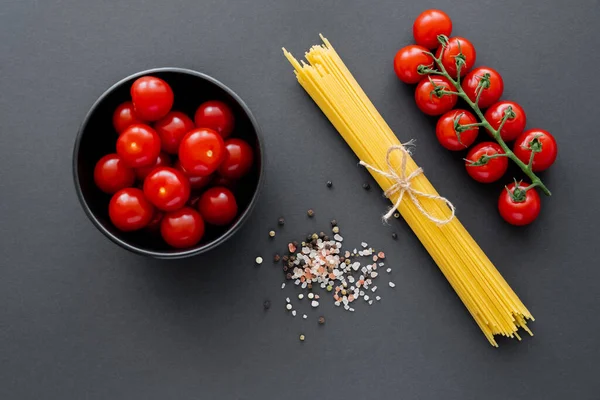 Draufsicht auf Kirschtomaten in Schüssel in der Nähe roher Pasta und Gewürze auf schwarzer Oberfläche — Stockfoto