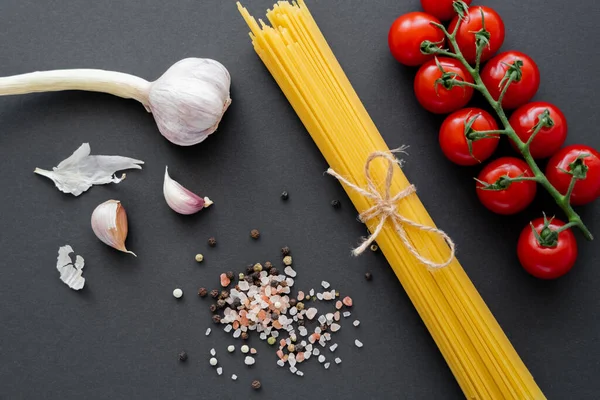 Vista dall'alto di aglio e pomodorini vicino spezie e pasta cruda su superficie nera — Foto stock