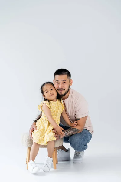 Повна довжина щасливої азіатської дівчини в літньому платті, що стоїть біля батька на сірому — стокове фото